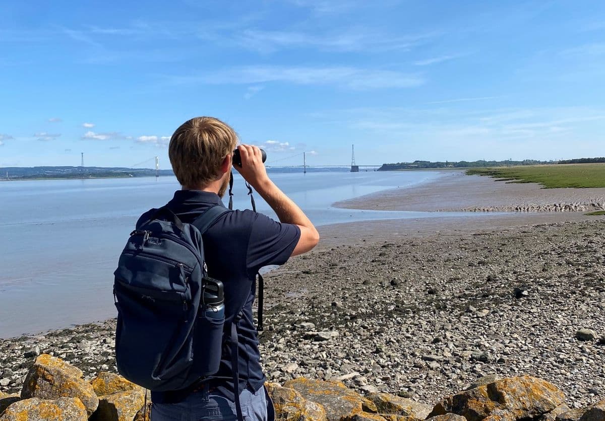 A man with a rucksack looking through binoculars at an estuary with a large bridge in the distance