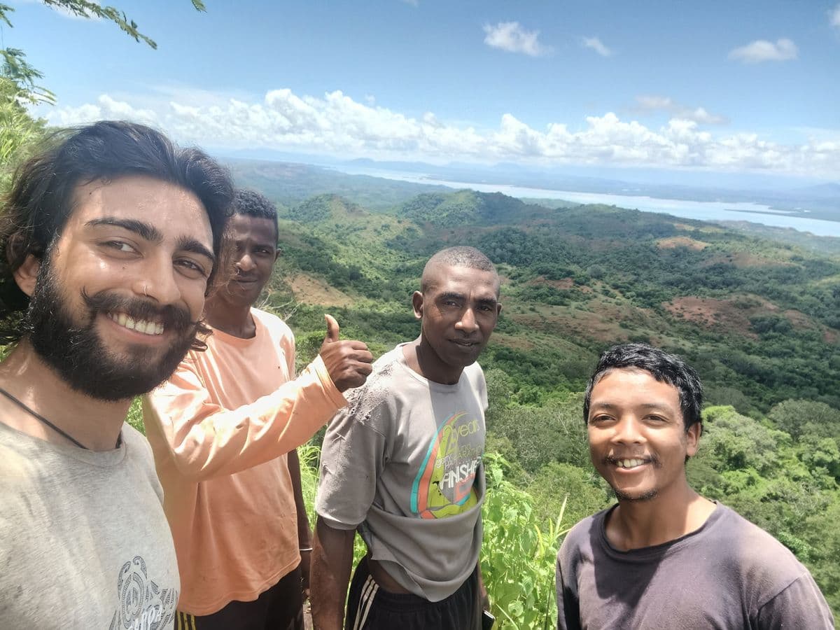 Four people smiling on a hilltop with a lush green landscape and distant water view in the background under a clear blue sky.